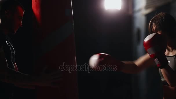 Tânăra frumoasă femeie brunetă în îmbrăcăminte sport se antrenează cu antrenorul său de box. Ţine pumni cu mănuşi de box în pungă pe fundal negru. Închidere — Videoclip de stoc