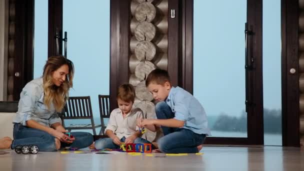Szczęśliwa matka z dwójką dzieci siedząc na podłodze jego domu kraju, spędzać czas razem zbierania projektanta dla dzieci. Pokój dzienny z panoramicznymi oknami. — Wideo stockowe