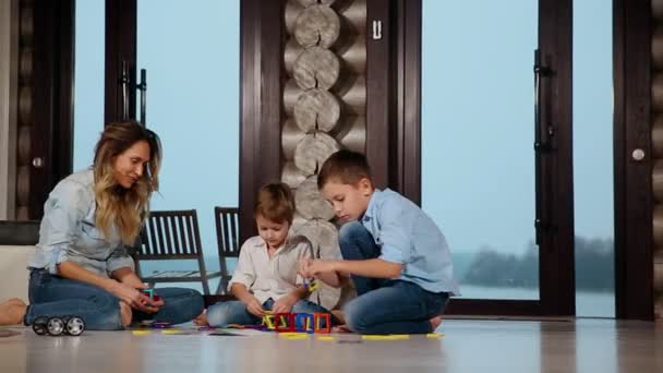 Madre feliz con dos niños sentados en el suelo de su casa de campo pasar tiempo juntos coleccionando diseñador de niños. Salón con ventanas panorámicas . — Vídeo de stock