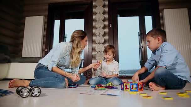 Gelukkig moeder met twee kinderen, zittend op de vloer van zijn landhuis tijd samen doorbrengen verzamelen childrens ontwerper. Woonkamer met panoramische ramen. — Stockvideo