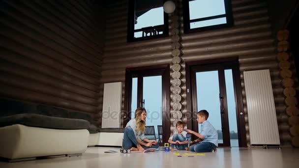 Gelukkig moeder met twee kinderen, zittend op de vloer van zijn landhuis tijd samen doorbrengen verzamelen childrens ontwerper. Woonkamer met panoramische ramen. — Stockvideo