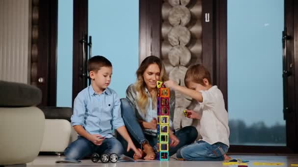 快乐的母亲与两个孩子坐在他的乡间别墅的地板上一起收集儿童设计师的时间。全景窗起居室. — 图库视频影像