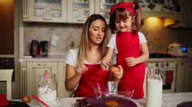 Anne ve kızı mutfakta bir kaseye yumurta kırma kırmızı önlük içinde bir pasta yapmak.