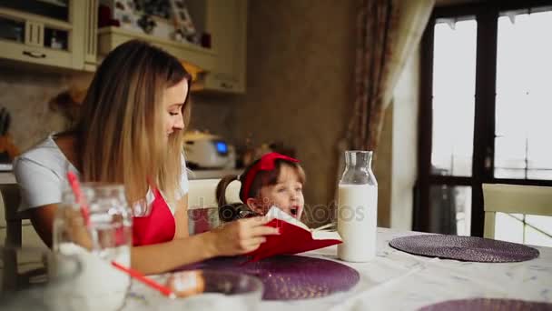 Μαμά και κόρη στην κουζίνα σε μια κόκκινη ποδιά Διαβάστε τη συνταγή κέικ σε ένα σημειωματάριο. — Αρχείο Βίντεο