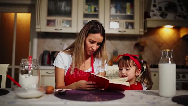 Mamma och dotter i köket i ett rött förkläde Läs recept kakan i en anteckningsbok. — Stockvideo