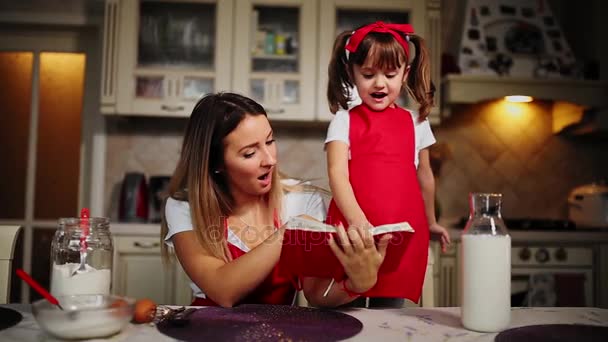 Mamma e figlia in cucina in grembiule rosso leggono la torta di ricette in un quaderno . — Video Stock