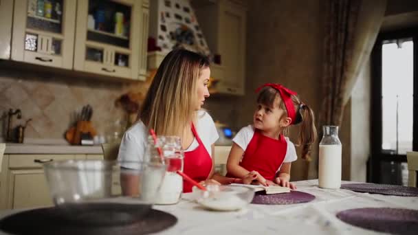 Μαμά και κόρη στην κουζίνα σε μια κόκκινη ποδιά Διαβάστε τη συνταγή κέικ σε ένα σημειωματάριο. — Αρχείο Βίντεο