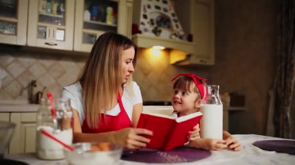 ママと赤エプロンで台所で娘を読んでノートにレシピ ケーキ. — ストック動画