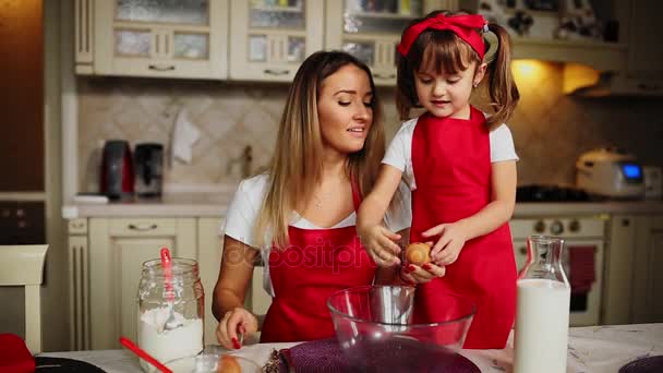 Anne ve kızı mutfakta bir kaseye yumurta kırma kırmızı önlük içinde bir pasta yapmak. — Stok video