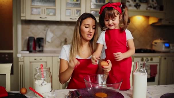 Μητέρα και κόρη στην κόκκινη ποδιές στην κουζίνα σπάζοντας τα αυγά σε ένα μπολ για να κάνει ένα κέικ. — Αρχείο Βίντεο