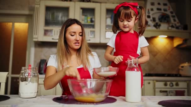 Vacker ung mamma att hjälpa sin lilla dotter tillsammans för att laga tårta i röda förkläden. Häll mjölet i en skål och vispa smeten för att göra en tårta i köket. — Stockvideo