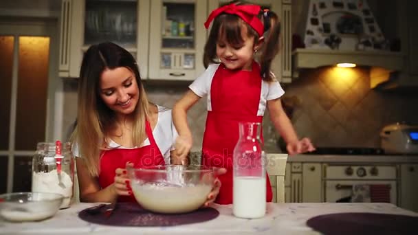 Die schöne junge Mutter hilft ihrer kleinen Tochter beim Kuchenbacken in roten Schürzen. Das Mehl in eine Schüssel geben und den Teig schlagen, um in der Küche einen Kuchen zu backen. — Stockvideo