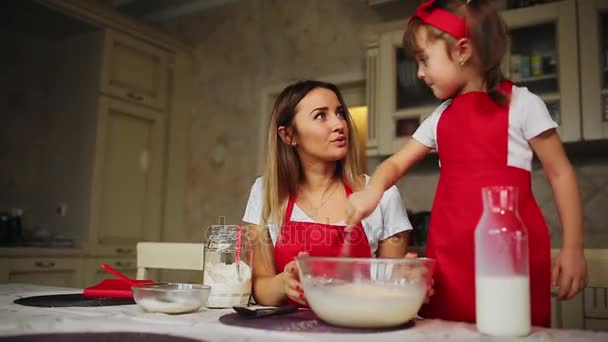 Bella giovane madre che aiuta la sua bambina a cucinare la torta in grembiuli rossi. Versare la farina in una ciotola e battere la pastella per fare una torta in cucina . — Video Stock