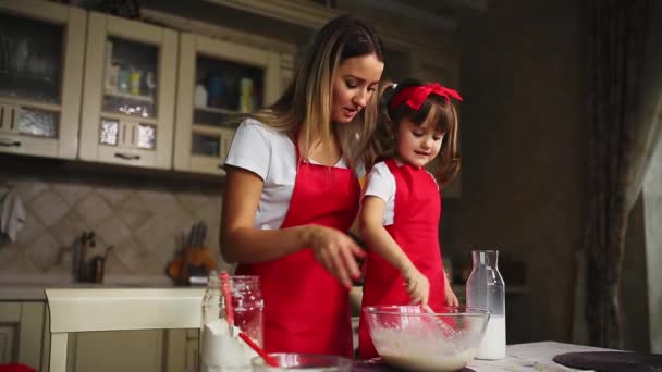 Όμορφη νεαρή μητέρα που βοηθούν τη μικρή της κόρη μαζί για να μαγειρέψουν το κέικ με κόκκινες ποδιές. Ρίξτε το αλεύρι σε ένα μπολ και χτύπησαν το κτύπημα για να κάνει ένα κέικ στην κουζίνα. — Αρχείο Βίντεο