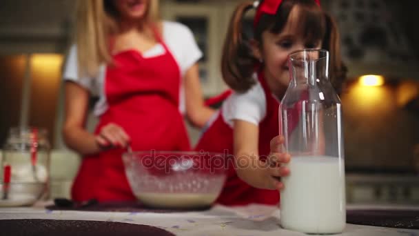 Hermosa madre joven ayudando a su hija pequeña a cocinar pastel en delantales rojos. Vierte la leche y revuelve la masa en la cocina. . — Vídeos de Stock
