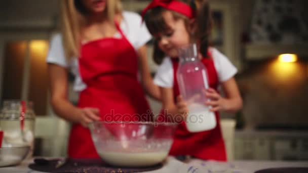 Όμορφη νεαρή μητέρα που βοηθούν τη μικρή της κόρη μαζί για να μαγειρέψουν το κέικ με κόκκινες ποδιές. Ρίχνουμε το γάλα και ανακατεύετε το χυλό μαζί στην κουζίνα. — Αρχείο Βίντεο