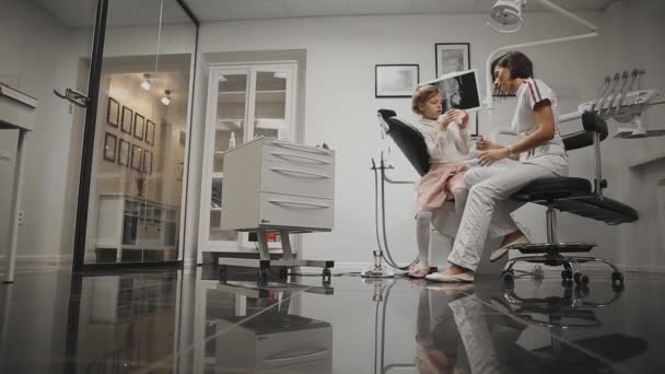 In de tandartsen office orthodontist op de achtergrond van de x-ray-hoofd van een klein meisje leren poetsen van je tanden na het installeren van bretels. De tandarts voert een raadpleging voor gebit reinigen. — Stockvideo