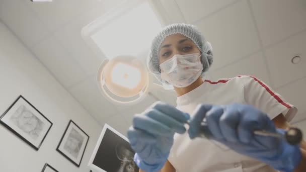 La joven dentista se inclina sobre el paciente gira la luz de la lámpara, trabajando con herramientas en máscara médica mirando a la cámara. La primera persona . — Vídeo de stock