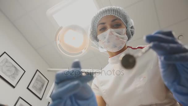 Молодая девушка-дантист наклоняется над пациентом, поворачивает лампу, работая с инструментами в медицинской маске, глядя в камеру. От первого лица . — стоковое видео