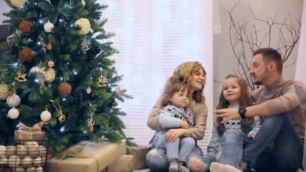 Famille heureuse en pull maman papa et deux filles assises sous le sapin à Noël, riant et s'achetant des chocolats . — Video