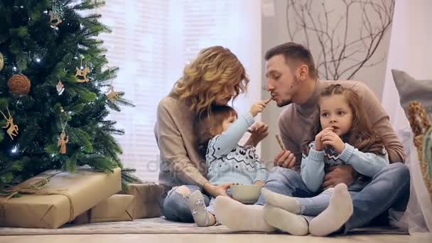 Gelukkige familie in truien moeder vader en twee dochters, zittend onder de boom met Kerstmis, lachen en elkaar chocolade kopen. — Stockvideo