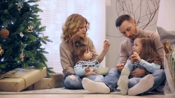 Famille heureuse en pull maman papa et deux filles assises sous le sapin à Noël, riant et s'achetant des chocolats . — Video