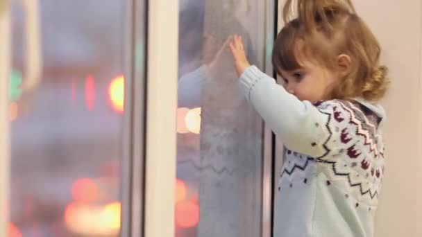 A little girl stands on the window sill in the winter and looks through the window at the passing cars. — Stock Video