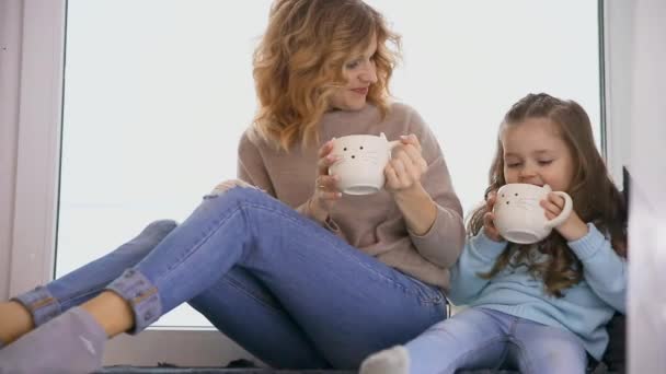 Madre e hija sentadas en el alféizar de la ventana en suéteres en Navidad y mirando por la ventana, hablar entre sí y beber café . — Vídeos de Stock