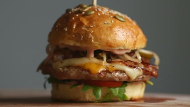 Stor hamburgare med nötkött kotlett, tomater, champinjoner och gurkor med smält ost roterar på en träskiva på ljus bakgrund. — Stockvideo