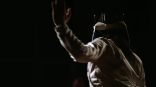 一个男人戴着一顶头盔, 在黑暗的背景下用剑杆与对手搏斗。围绕轴的运动和焦点在后面的敌人后视图 . — 图库视频影像