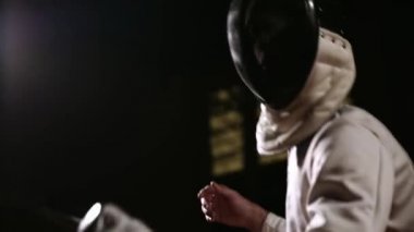 Bir adam closeup bir rapier ile karanlık bir arka plan üzerinde bir rakip ile kıskaç eskrim için kask takıyor. Kameranın alt kısmından yukarı taşır.
