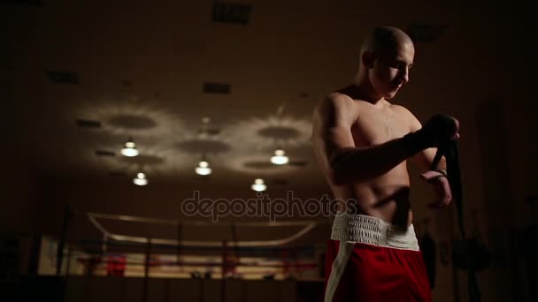 Ein aufgepumpter Mann in Boxstiefeln und kurzen Hosen in der Turnhalle vor dem Hintergrund eines Boxrings trägt Bandagen an den Händen. — Stockvideo