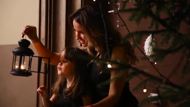 Mamá y su hija se sientan bajo un árbol de Navidad apagan una antorcha con una vela . — Vídeo de stock