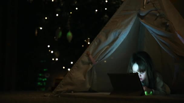 Daheim in einem Zelt vor dem Hintergrund eines Weihnachtsbaums sitzend, lernt ein kleines Mädchen, das auf dem Boden liegt und über Videokommunikation spricht, den Umgang mit einem Tablet-Computer. Nahaufnahme — Stockvideo