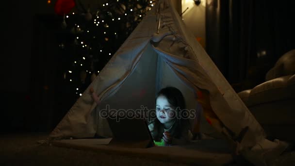 Sentado en casa en una tienda de campaña sobre el fondo de un árbol de Navidad una niña pequeña acostada en el suelo examina su nuevo regalo, aprende a usar una tableta — Vídeos de Stock