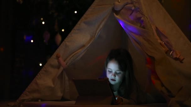 Sentado en casa en una tienda de campaña sobre el fondo de un árbol de Navidad, una niña tendida en el suelo entra en una consulta de búsqueda en una tableta electrónica . — Vídeos de Stock