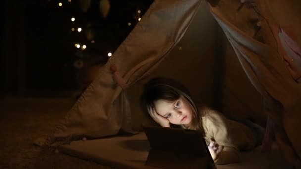 Om thuis te zitten in een tent tegen de achtergrond van een kerstboom, een klein meisje op de vloer liggen houdt op haar hoofd, en met de andere hand persen ze het scherm van de tablet van de elektronische. — Stockvideo