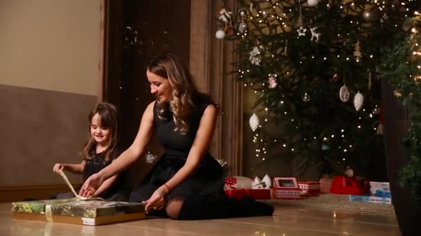 Sentada bajo un árbol de Navidad en la víspera de Navidad, mi madre le dio a su hija un regalo y una chica desempacó cintas de regalo atadas. Movimiento de cámara — Vídeos de Stock