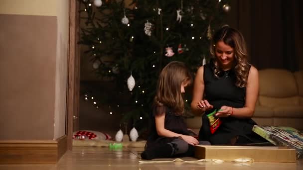 Anya és lánya a karácsonyfa alatt ül megnyitni ajándékok. Egy lány, egy ajándék szalag függetlenítése — Stock videók
