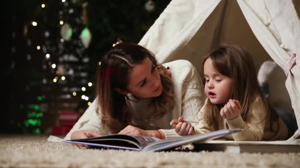 Vestidos con suéteres blancos, la madre y la hija leen un cuento de hadas para la noche acostadas con ella en la tienda de una casa establecida en el fondo de un árbol de Navidad — Vídeos de Stock
