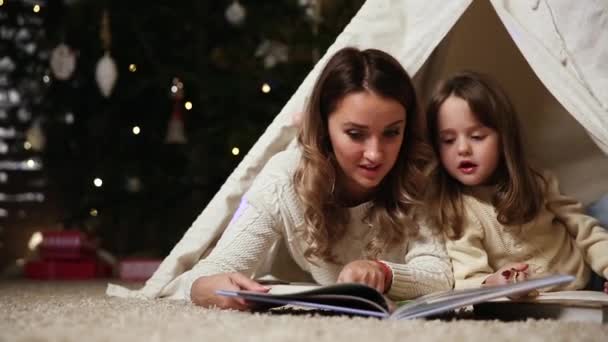 Anya olvasó egy lánya mese az éjszaka, fekvő vele a sátorban, egy megalapozott ház a karácsonyfa háttér — Stock videók