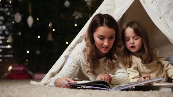 Mamá está leyendo un cuento de hijas para la noche acostada con ella en la tienda de una casa establecida en el fondo de un árbol de Navidad — Vídeos de Stock