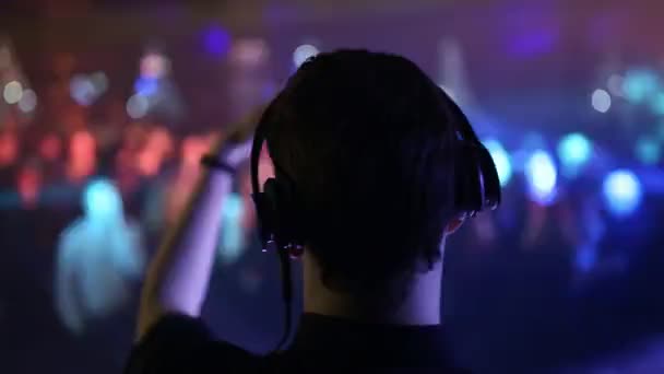 Vista traseira. As costas e fones de ouvido do DJ em primeiro plano, as pessoas que dançam são borradas . — Vídeo de Stock