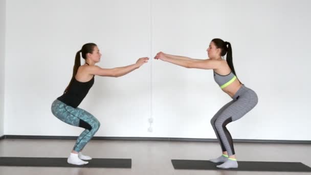 Duas meninas em roupas esportivas em um estúdio em um fundo branco executar agachamentos uns contra os outros. Enchimento simultâneo. Treinamento composto — Vídeo de Stock