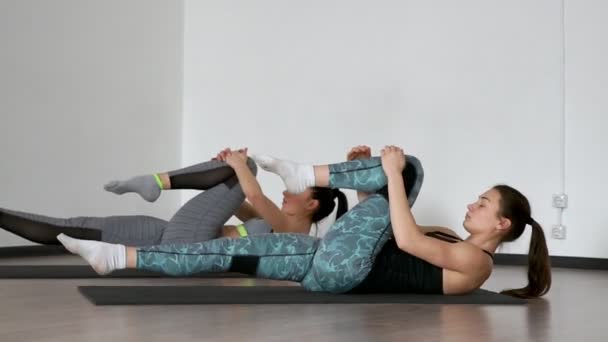 Im Fitnessstudio während der Pilates-Übung auf den Gymnastikmatten liegend ziehen zwei Mädchen, die auf dem Rücken liegen, abwechselnd ihre Knie zur Brust und dehnen die Muskeln der Oberschenkel. synchrone Ausführung der — Stockvideo