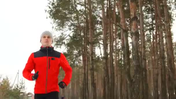 Un corredor profesional con una chaqueta roja pasa su entrenamiento en el aire fresco corriendo por el bosque de pinos en invierno — Vídeos de Stock