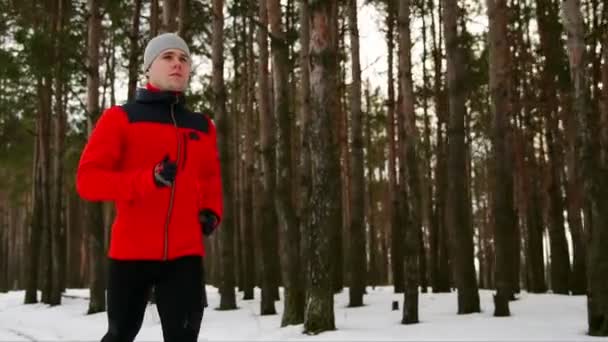 A young sporty man in a red jacket spends his training in the fresh air running along the pine forest in winter. Slow motion. 120 frames per second — Stock Video