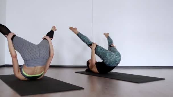 Twee meisjes in de sportschool uitvoeren een oefening klubochalka Pilatus hassel. Lezhnaya op haar rug met haar benen verhoogd maken een roll op haar rug. de camera gaat in een baan. Doen oefening op een wit — Stockvideo