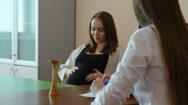 Siyah T-shirt genç bir güzel hamile kadın genç kadın doktor hastanede ofisinde beyaz paltolu konuşuyor. Doktor ve hasta Danışma