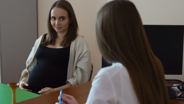 Молодая красивая беременная женщина в черной футболке разговаривает с молодой докторшей в белом халате в своем кабинете в больнице — стоковое видео